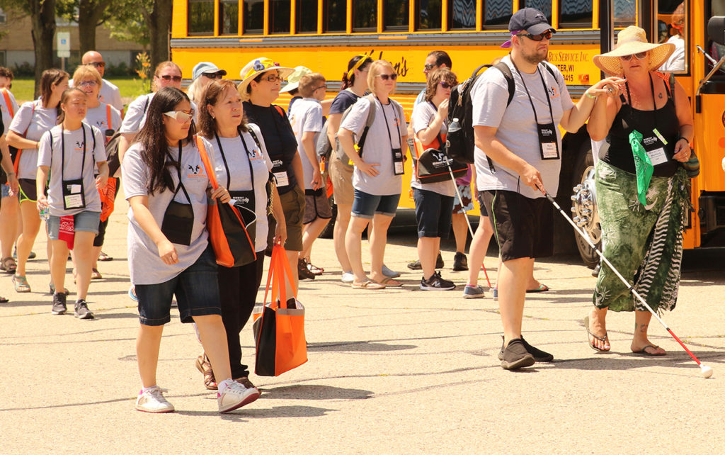 Large group of people walking past the camera. One person is using a white cane and is holding the shoulder of another person. There is a school bus in the background.