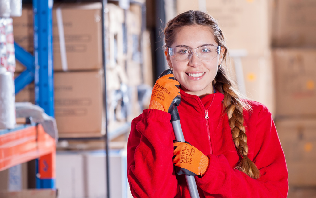 Young girl with long hair in a braid draped over her shoulder. She is holding a broom handle and wearing a red zip-up fleece jacket, orange work gloves, and clear safety glasses. In the background are heavy duty shelves lined with brown boxes.