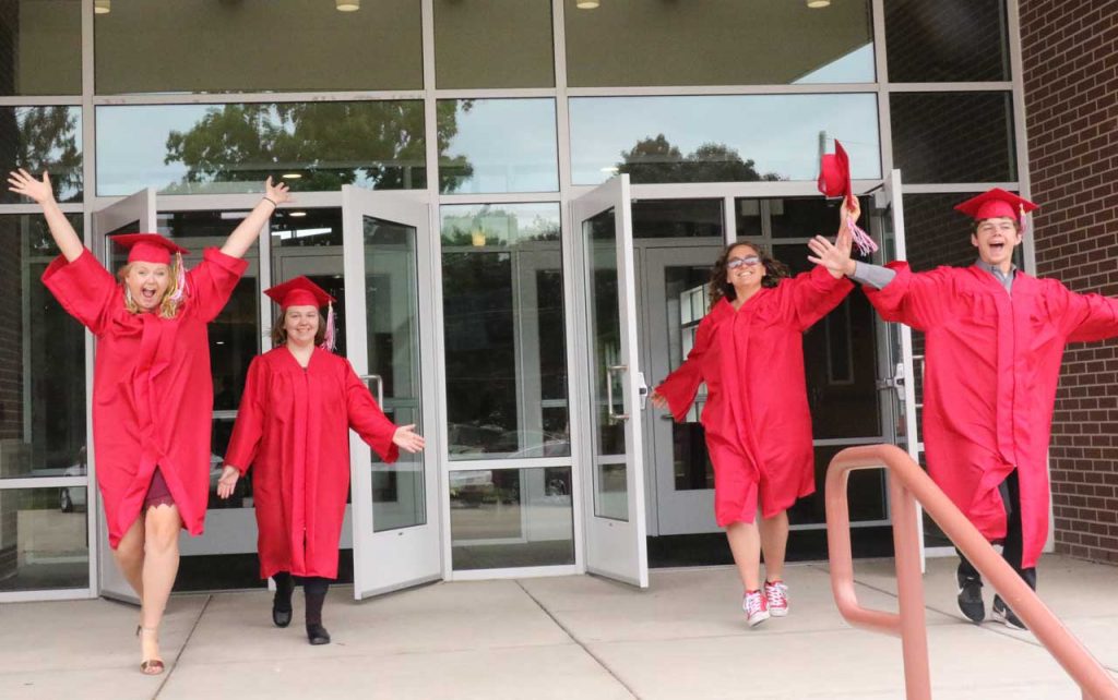 Three female and one male student wearing red graduation caps and gowns, exiting the school building in celebration.