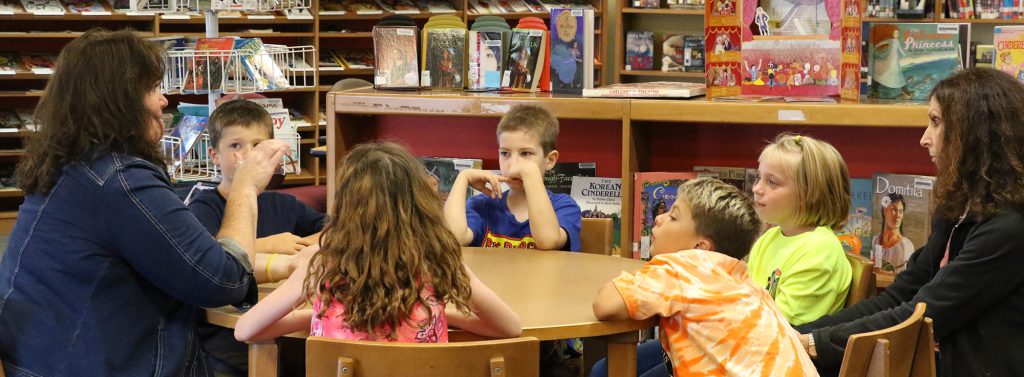 Elementary students sitting at a library table with teacher and educational assistant.