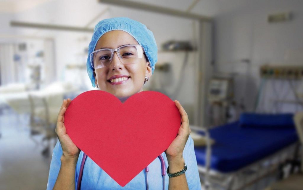 Nurse wearing a hairnet and safety glasses, holding up a red heart in front of her.