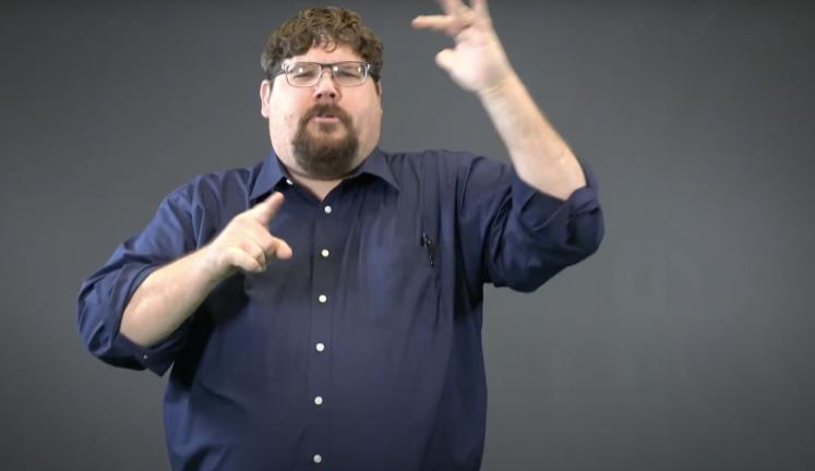 Man with dark hair, glasses, and a goatee wearing a dark blue shirt is using American Sign Language, explaining something to the camera.