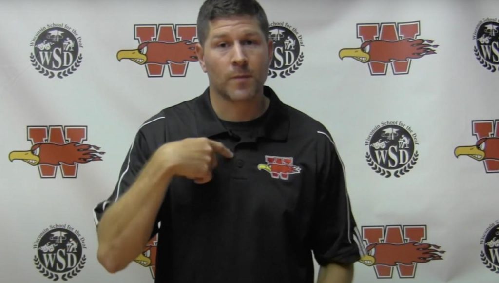 Man with dark hair wearing a black shirt with the WSD Firebird logo, standing in front of a white screen with the WSD school and athletic logos, using American Sign Language to explain something to the camera.