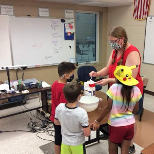 Three students standing around a table with their teacher, measuring flour into a mixing bowl.