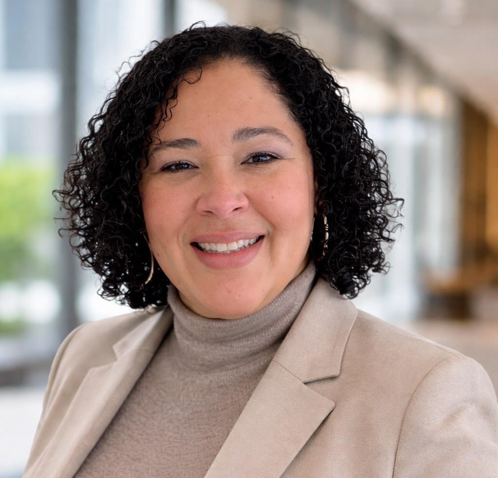 Professional headshot of a smiling woman with curly hair, wearing a beige blazer and turtleneck.