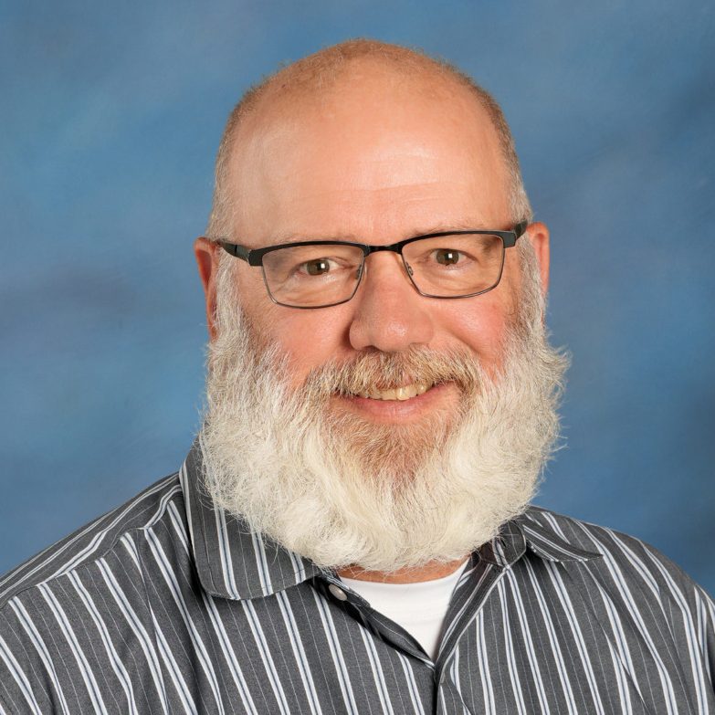 Professional headshot of a smiling bald man with a white beard and glasses, wearing a striped shirt against a blue background.
