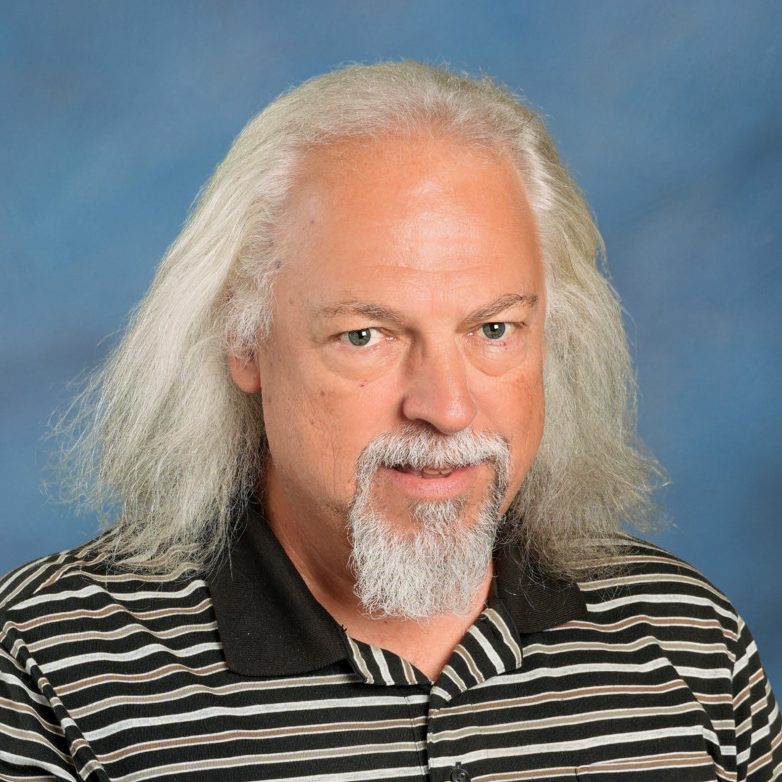 Professional headshot of a smiling man with a white hair and beard, wearing a striped shirt against a blue background.