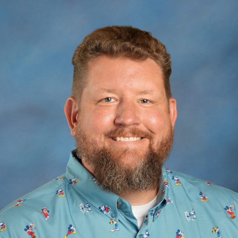 Professional headshot of a smiling man with a brown hair and beard, wearing a blue shirt against a blue background.