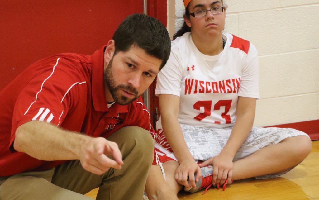 Male basketball coach pointing toward the camera with female player in the background.