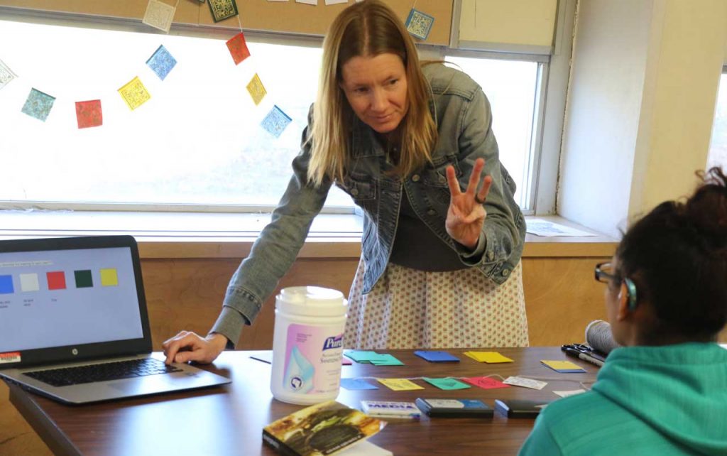 Female teacher with mid-length light brown hair demonstrating something on a Chromebook and holding up the number six in American Sign Language. A female student wearing a hearing aid sits at the table facing the teacher with her back to the camera. The table has colored paper and stamping supplies on it. A banner with prayer flags is displayed in the window behind the teacher.