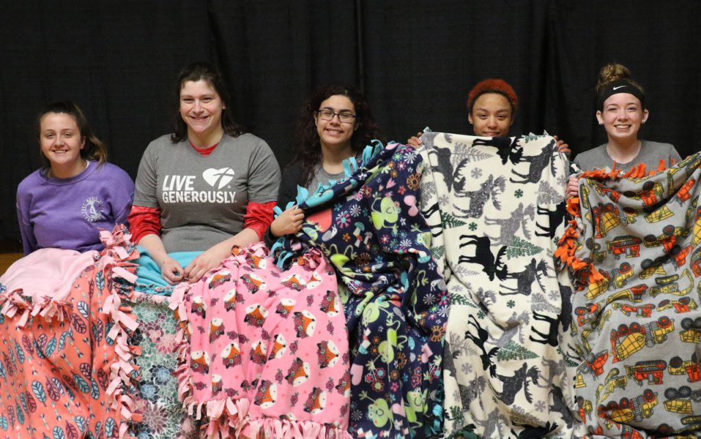 Five students sitting on a stage, holding up blankets they made.