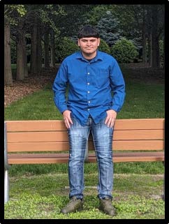 Nestor, a 2024 graduate, is wearing a navy blue button-up shirt while standing in front of bench in the park for his Senior photo
